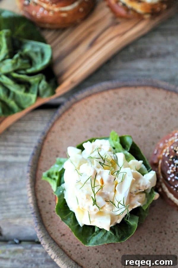 Dill Pickle Egg Salad is classic creamy egg salad packed with cubes of crispy dill pickles, and fresh dill. Served on a pretzel roll it's heaven! Dill Pickle Egg Salad on lettuce and pretzel roll, brown speckled pottery plate, lettuce leaves, wooden cutting board, rustic wood table, vertical