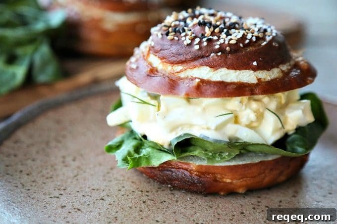 Dill Pickle Egg Salad is classic creamy egg salad packed with cubes of crispy dill pickles, and fresh dill. Served on a pretzel roll it's heaven! Dill Pickle Egg Salad on lettuce and pretzel roll, brown speckled pottery plate, lettuce leaves, wooden cutting board, rustic wood table, close up