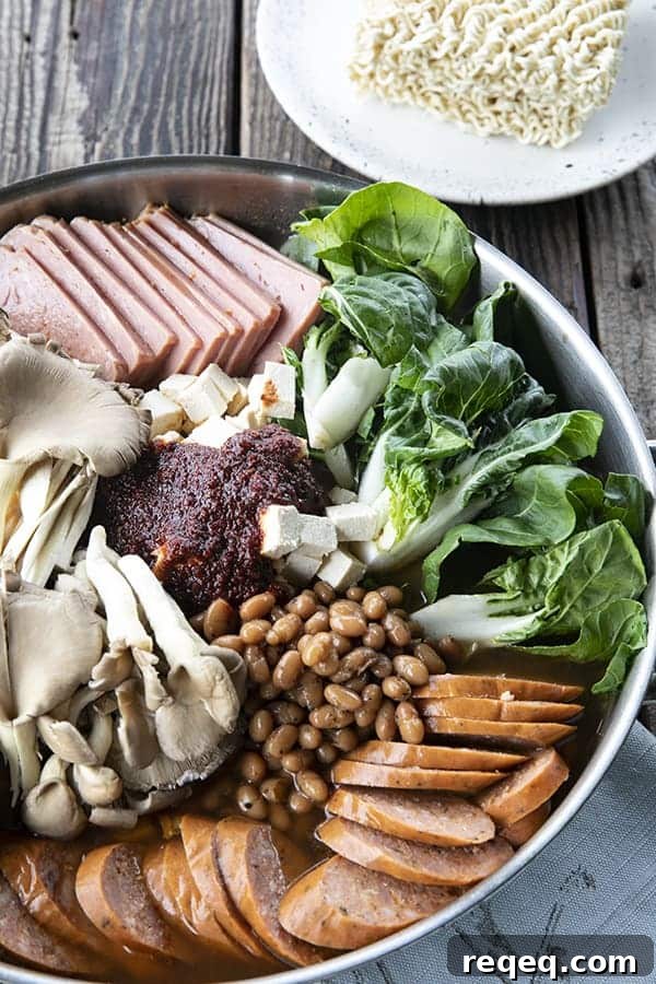Budae Jjigae (Korean Army Stew): Spicy Sausage Hot Pot 3 Korean Army Stew or Budae Jjigae in a stainless steel everyday pan, ready to simmer. Bok choy, sliced sausage, sliced spam, mushrooms, baked beans, gochujang seasoning, and ramen noodles on the side on a plate.