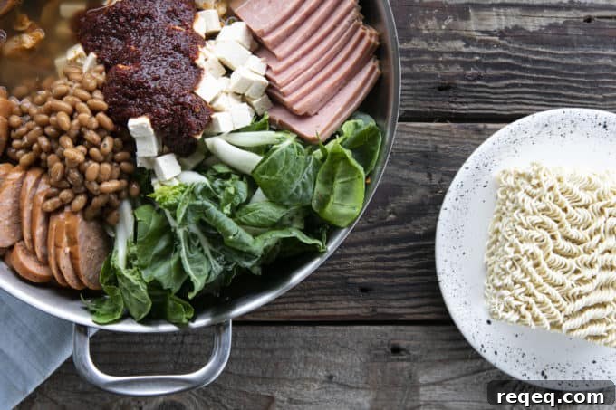 Budae Jjigae (Korean Army Stew): Spicy Sausage Hot Pot 4 Budae Jjigae or Korean Army Stew ready to simmer. Stainless steel everyday pan filled with bok choy, sliced sausace, spam, mushrooms, tofu cubes, baked beans, and sauce. Ramen noodles on a plate next to the pan.