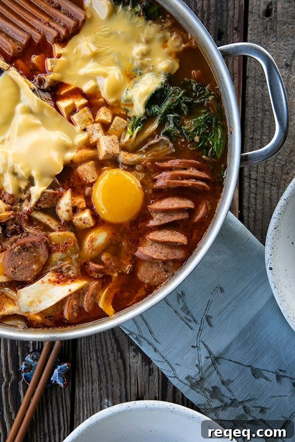 Budae Jjigae (Korean Army Stew): Spicy Sausage Hot Pot 5 Korean Army Stew or Budae Jjigae with egg yolks and American cheese in a stainless steel everyday pan. Blue linen on the table with two white bowls and wooden chopsticks.