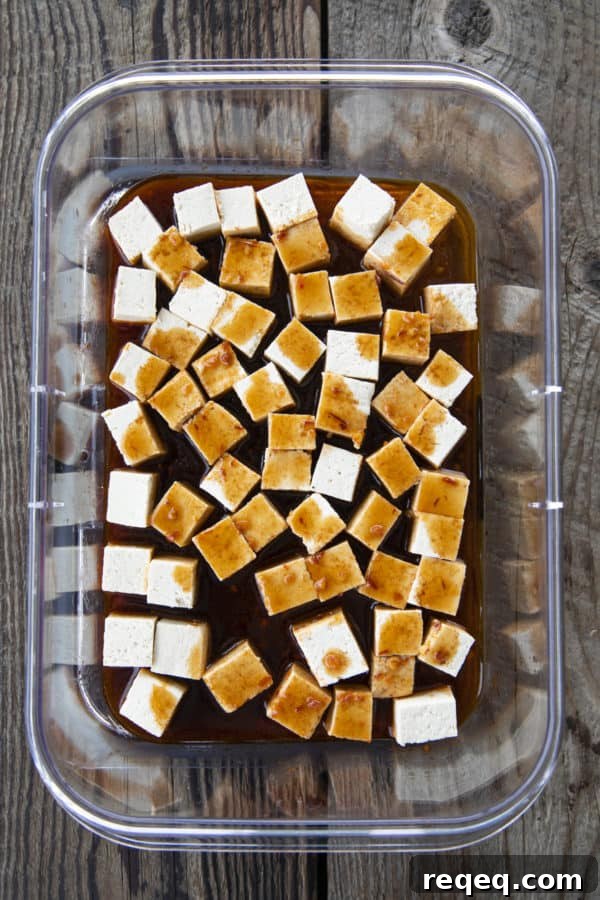 Honey Sriracha Crispy Tofu in the Air Fryer: Sticky & Crunchy 4 Small cubes of firm tofu in marinade in a transparent, rectangular container.