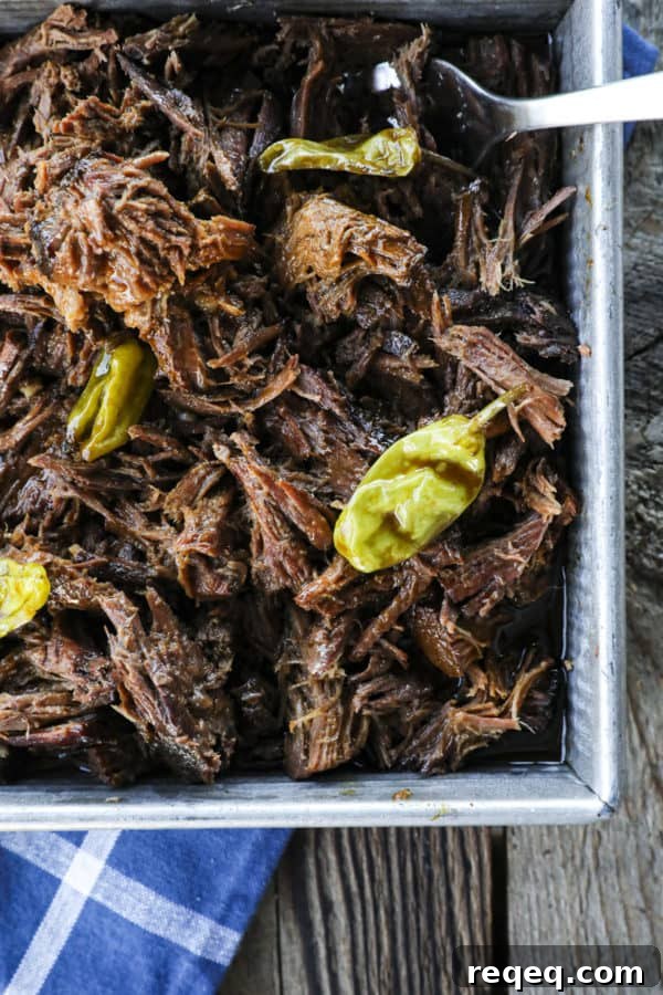 Mississippi Pot Roast Slow Cooker Recipe: Tender, Flavorful Roast 4 Shredded Crock Pot Mississippi Pot Roast in a square metal pan with whole pepperoncini and meat forks on a blue linen and wooden table.