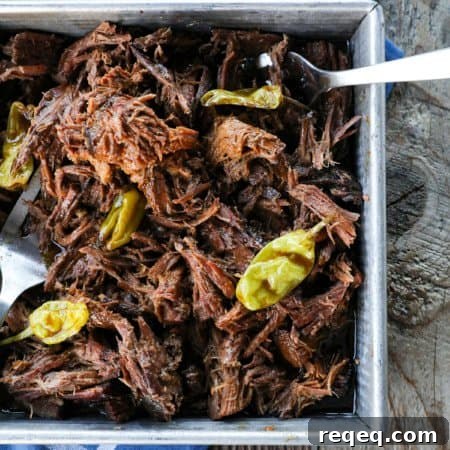 Mississippi Pot Roast Slow Cooker Recipe: Tender, Flavorful Roast 8 Shredded Crock Pot Mississippi Pot Roast in a square metal pan with whole pepperoncini and meat forks on a blue linen and wooden table.
