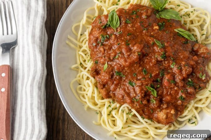 Slow-Cooker Spaghetti Sauce Recipe for Rich, Hands-Off Flavor 5 spaghetti sauce on spagetti on a plate topped by fresh herbs with a napkin and fork on the left.