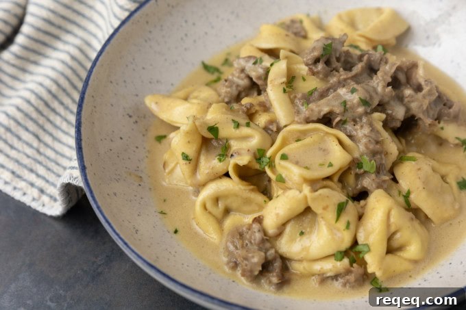 Philly cheesesteak tortellini in rich provolone sauce garnished with chopped parsley in a blue rimmed speckled bowl on a table with a blue and white striped linen