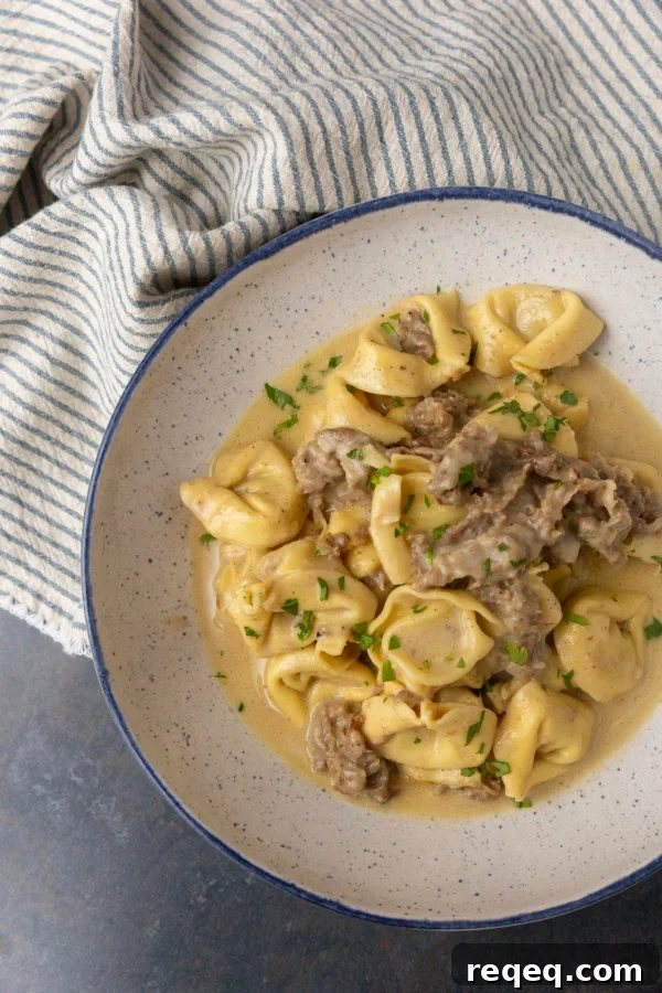 Philly cheesesteak tortellini in rich provolone sauce garnished with chopped parsley in a blue rimmed speckled bowl on a table with a blue and white striped linen