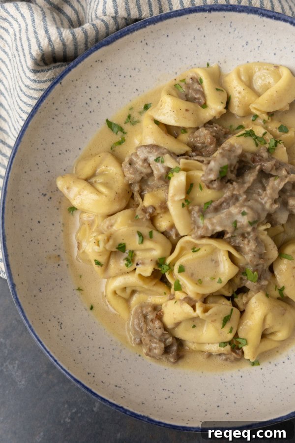 Philly cheesesteak tortellini in rich provolone sauce garnished with chopped parsley in a blue rimmed speckled bowl on a table with a blue and white striped linen