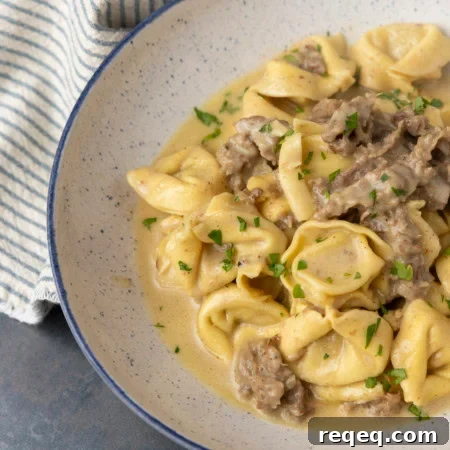 Philly cheesesteak tortellini in rich provolone sauce garnished with chopped parsley in a blue rimmed speckled bowl on a table with a blue and white striped linen